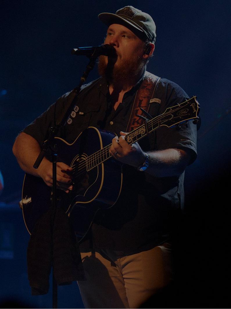 Man with beard wearing cap and denim shirt performing with acoustic guitar and microphone on stage.