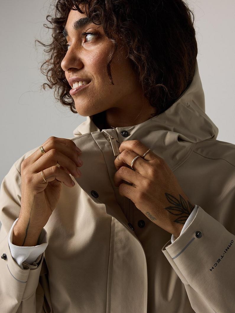 Person with curly hair smiling while adjusting the collar of a beige jacket, wearing gold rings.
