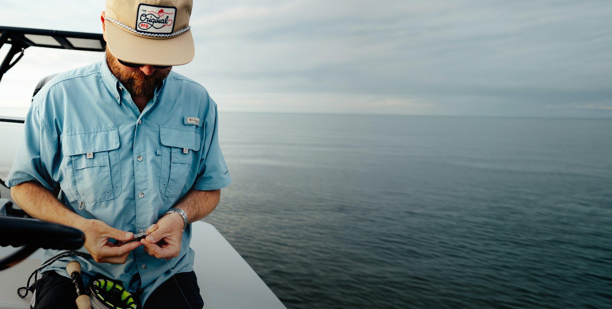 Man wearing light blue fishing shirt with multiple chest pockets, tying a fishing lure on a boat.