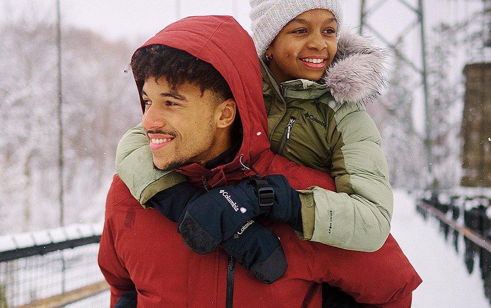 Un homme vêtu d'une veste rouge porte sur son dos un enfant qui porte des mitaines Columbia Sportswear.