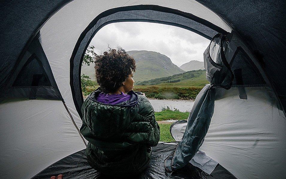 A person in a Columbia puffer coat sits in a tent looking out on a rain soaked valley and stream.