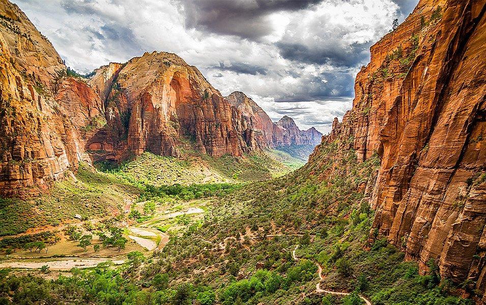 A canyon of orange and red rock stands on either side of the greenery and trail-filled land below.  