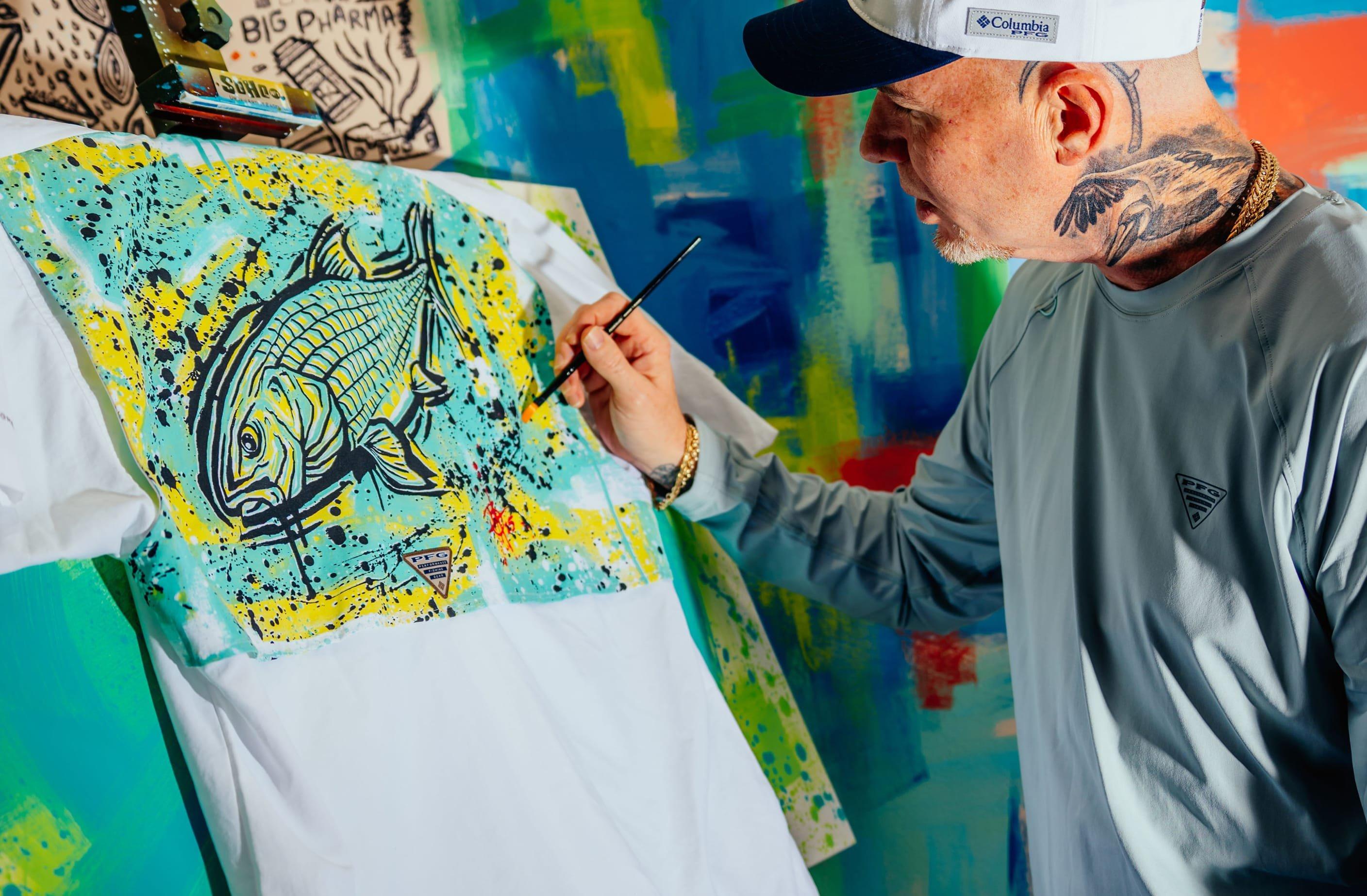 Artist painting a colorful fish design on a white performance fishing shirt using a brush.