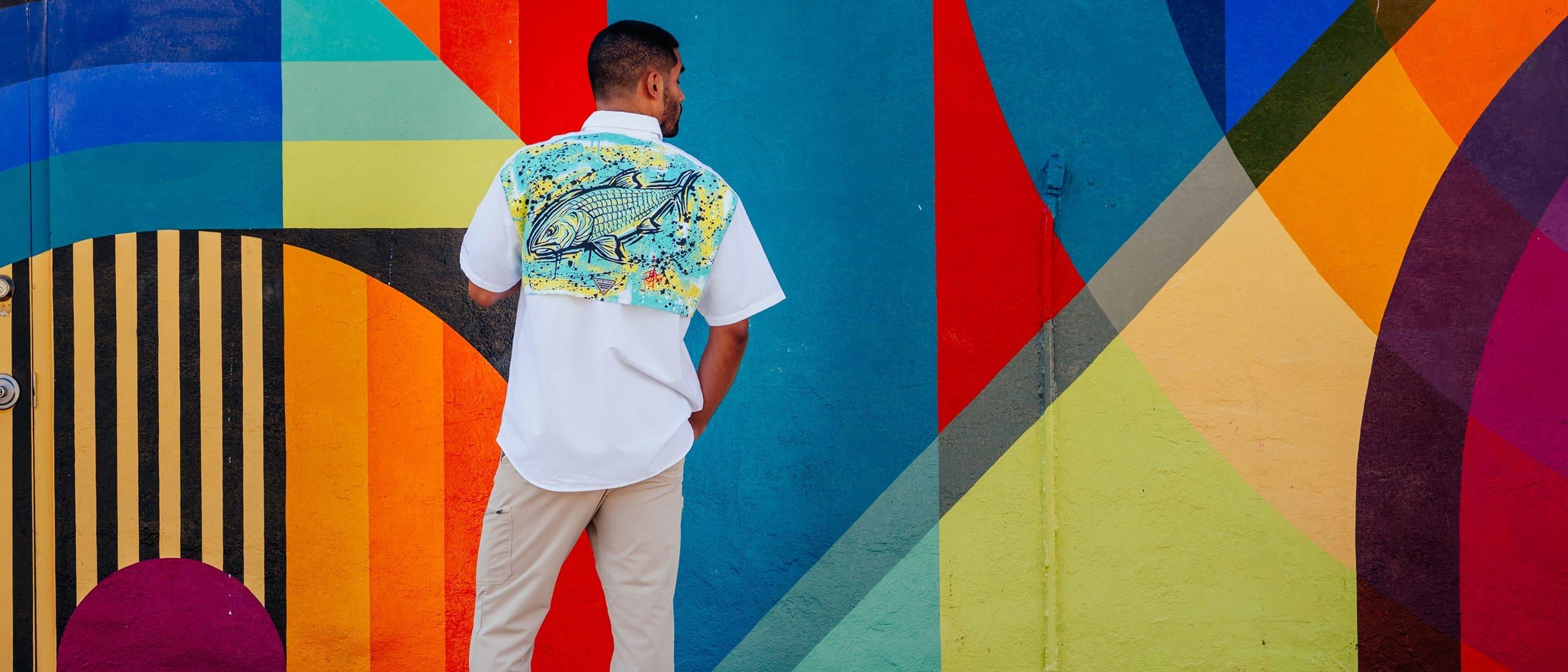 Man wearing white short-sleeve shirt with fish graphic on back, standing against colorful geometric mural.