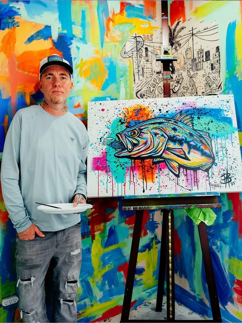 Artist standing beside easel displaying vibrant abstract fish painting with colorful splatter effects.
