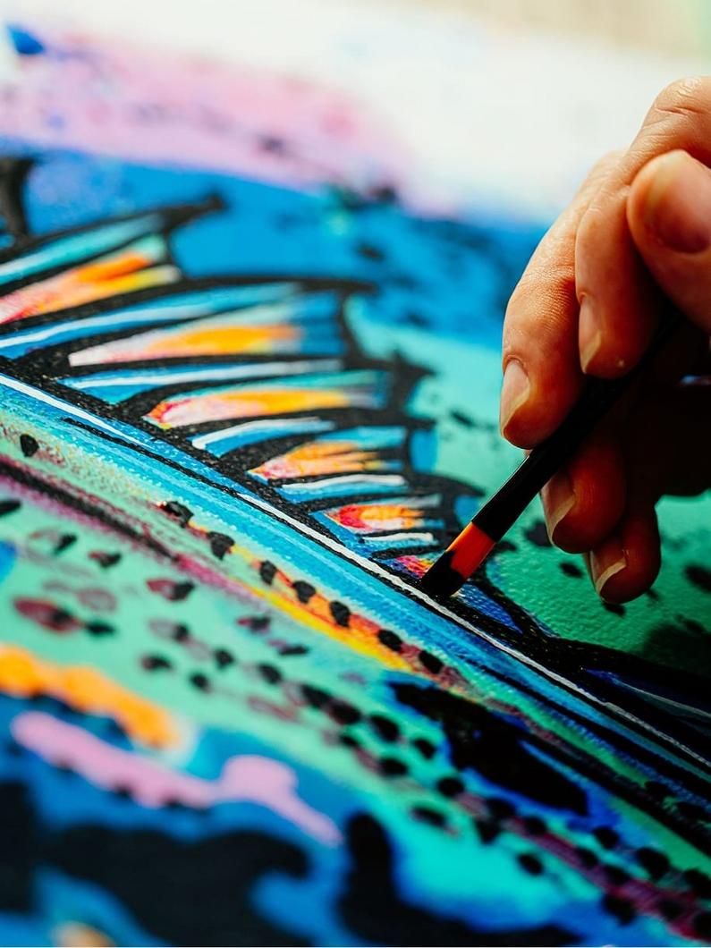 Hand holding paintbrush applying colorful strokes to an abstract canvas with blue, pink, and yellow hues.