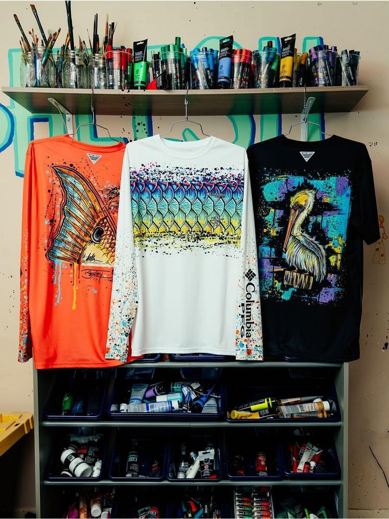 Three graphic t-shirts displayed on hangers featuring fish, geometric pattern, and pelican designs.