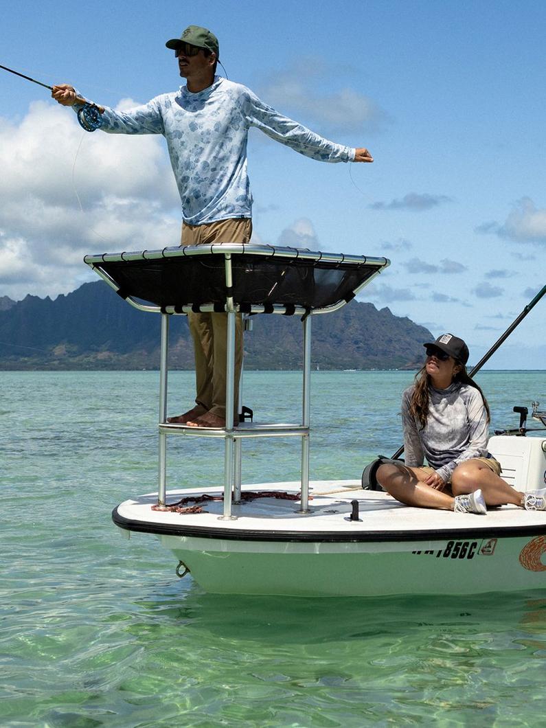 Man standing on boat platform holding fishing rod with woman sitting on deck in tropical waters.