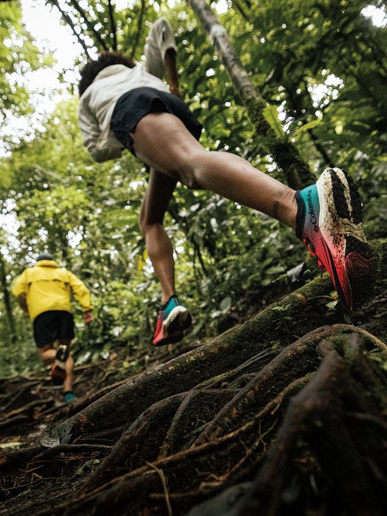 Person leaping over mossy log on forest trail wearing colorful athletic shoes with another runner behind.