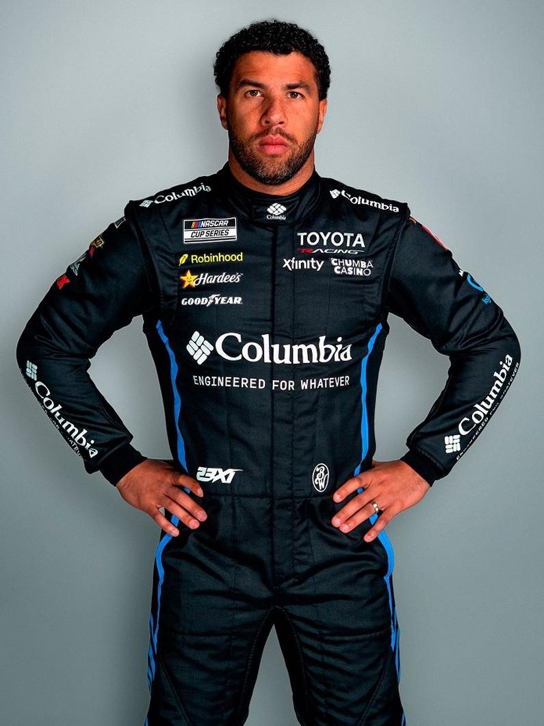 Race car driver posing in black racing suit with hands on hips and sponsor logos.