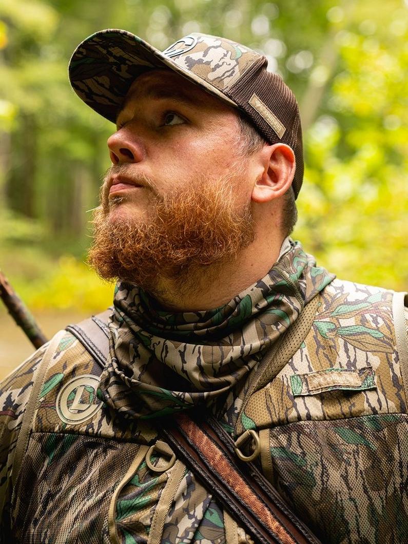 Man wearing camouflage hunting apparel with neck gaiter and cap, looking upward in outdoor setting.