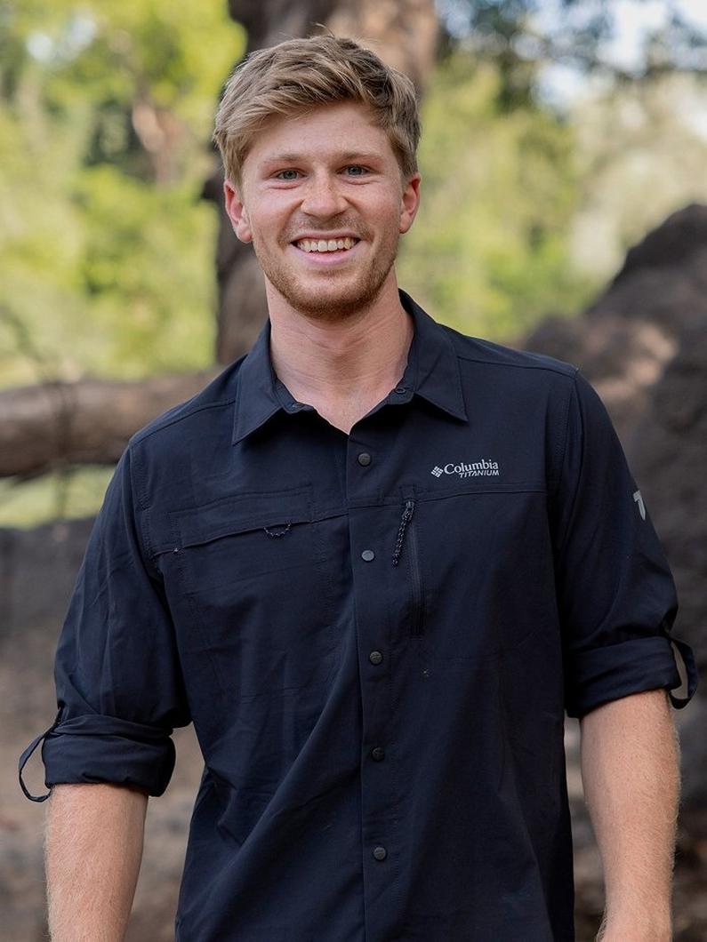 Man smiling outdoors wearing a navy button-up shirt with rolled sleeves and chest pockets.