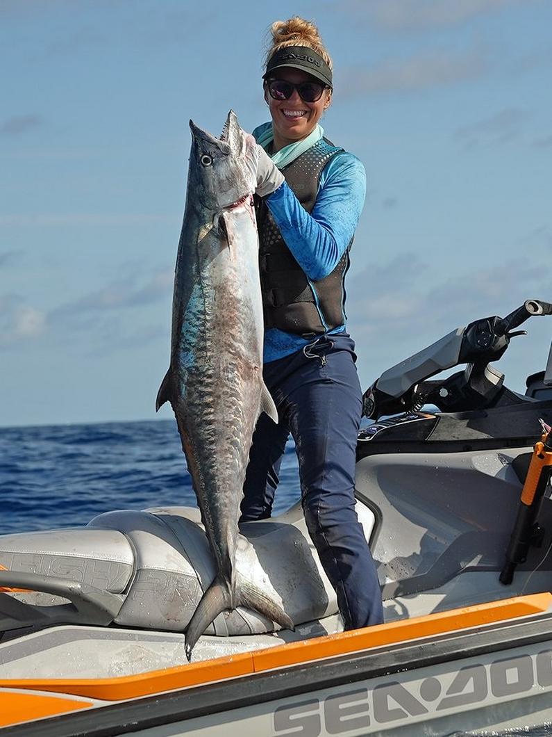 Woman proudly holding a large caught fish while standing on a watercraft in the ocean.