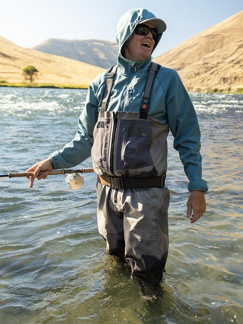 Man wearing teal jacket and waders standing in river holding fishing rod with reel.