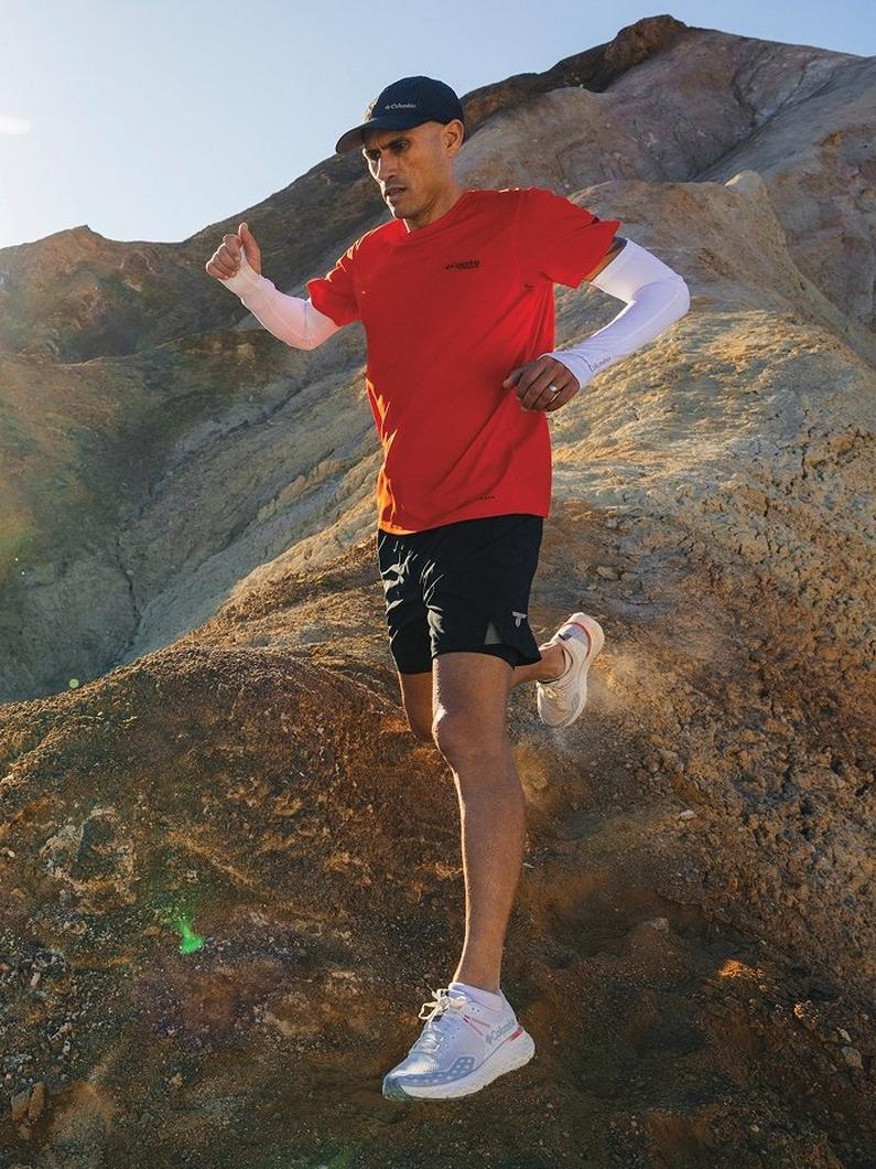 Man running on rocky mountain terrain wearing red t-shirt, black shorts, white sneakers, and cap.