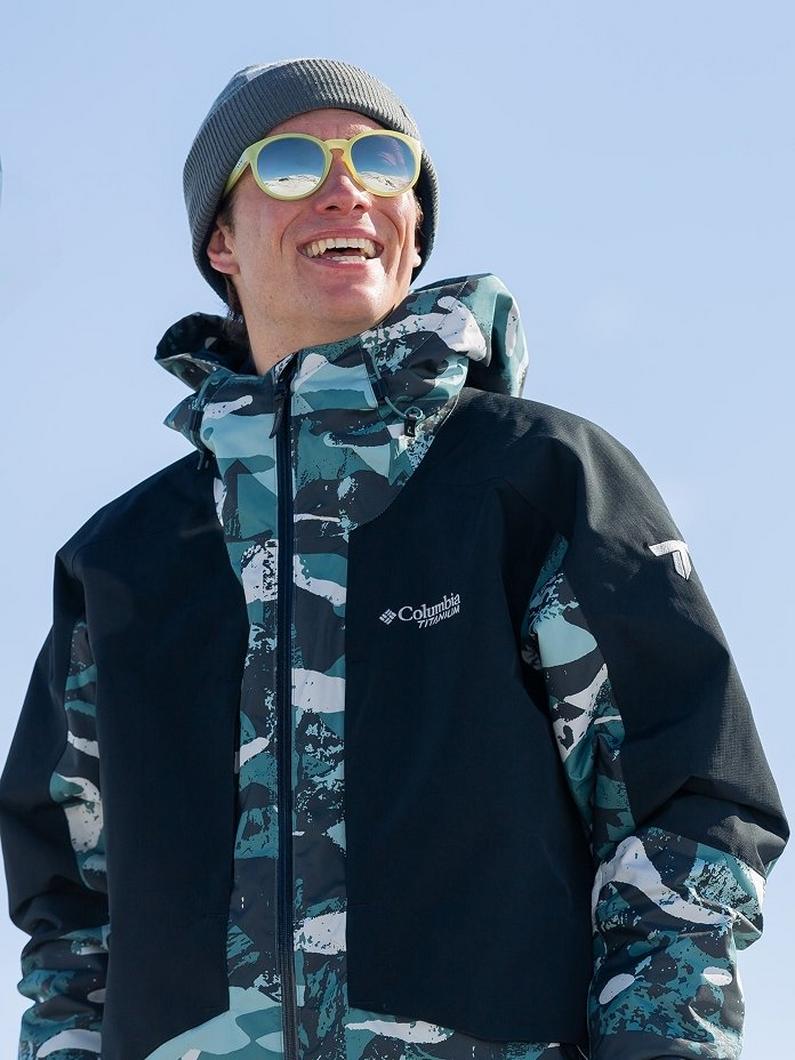 Man smiling outdoors wearing gray beanie, yellow sunglasses, and black jacket with teal camo accents.