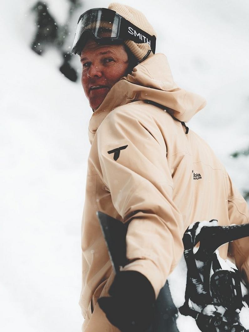 Person wearing beige ski jacket, beanie, and goggles while holding a snowboard in snowy conditions.