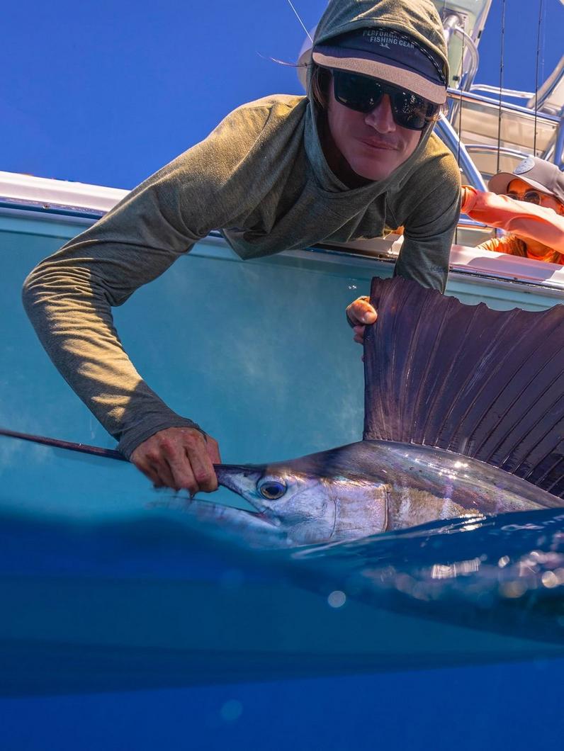 Angler in sunglasses and cap holding a sailfish in the water beside a boat.