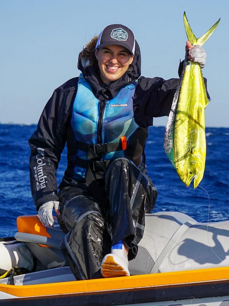 Woman on jet ski holding up a large yellow mahi-mahi fish while wearing a life jacket and cap.