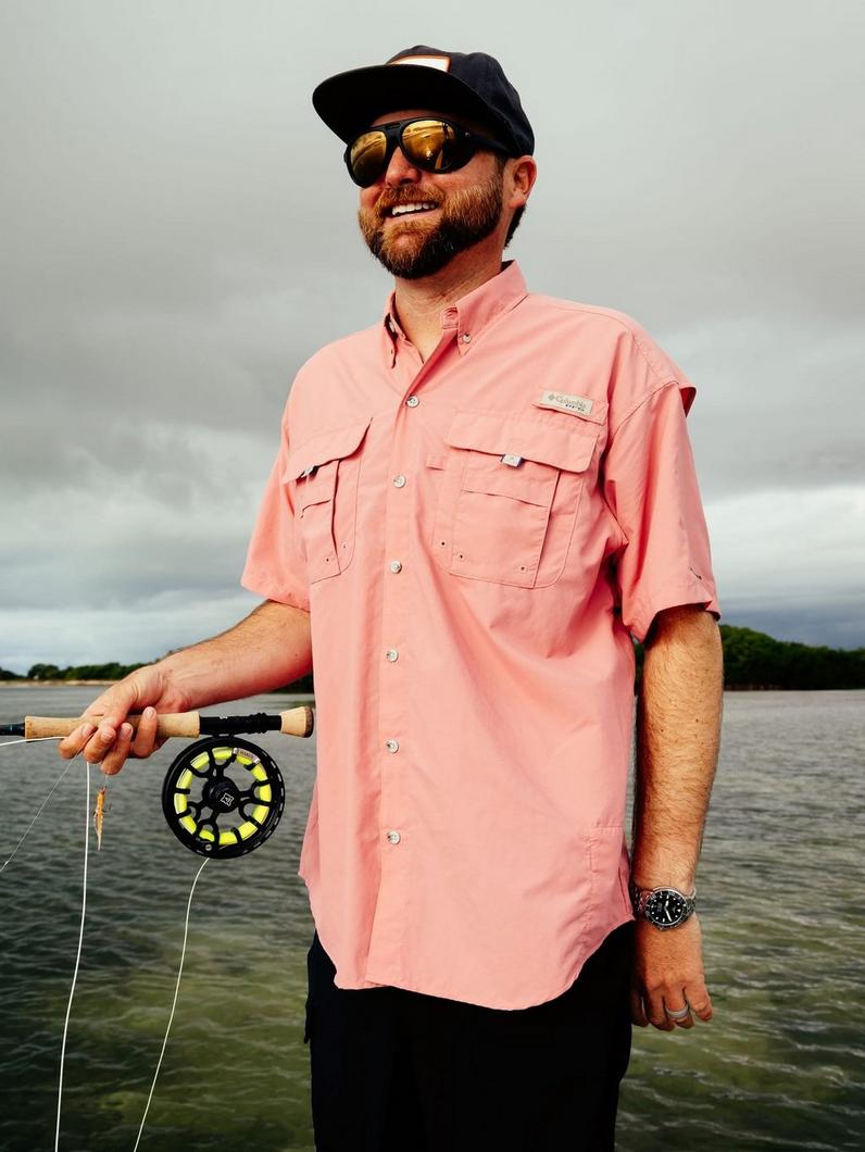 Man wearing coral fishing shirt and sunglasses holding fishing rod by water.