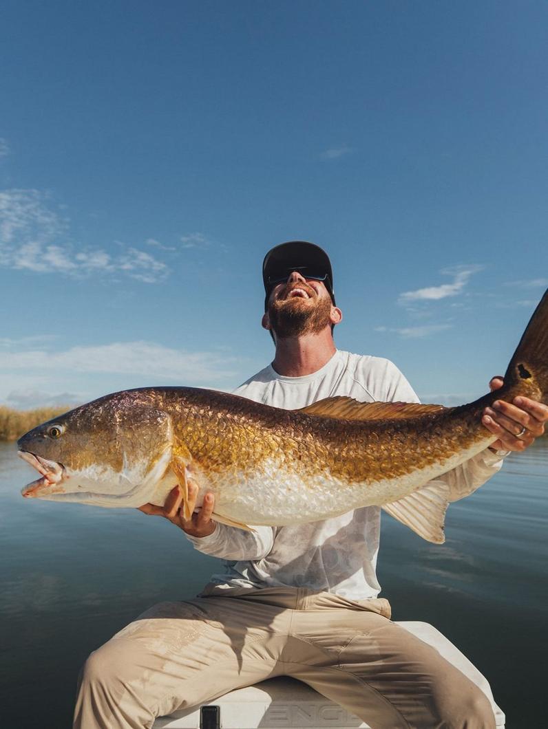 Man in white shirt and cap proudly holding large redfish while sitting on boat under blue sky.