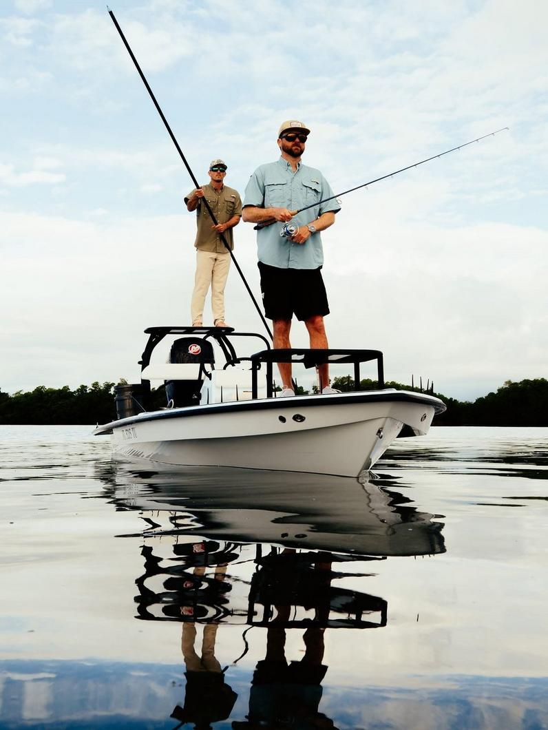 Two men fishing from a white boat on calm water, wearing fishing shirts, caps, and sunglasses.