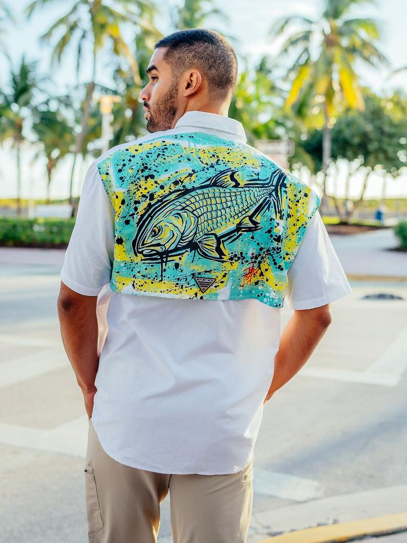 White short-sleeve shirt with turquoise and yellow fish graphic on back, modeled outdoors near palm trees.