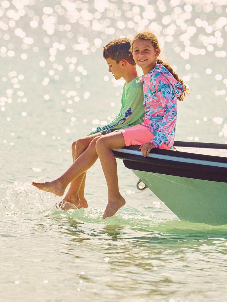 Two children sitting on the edge of a boat with their feet in the water, wearing colorful swim shirts.
