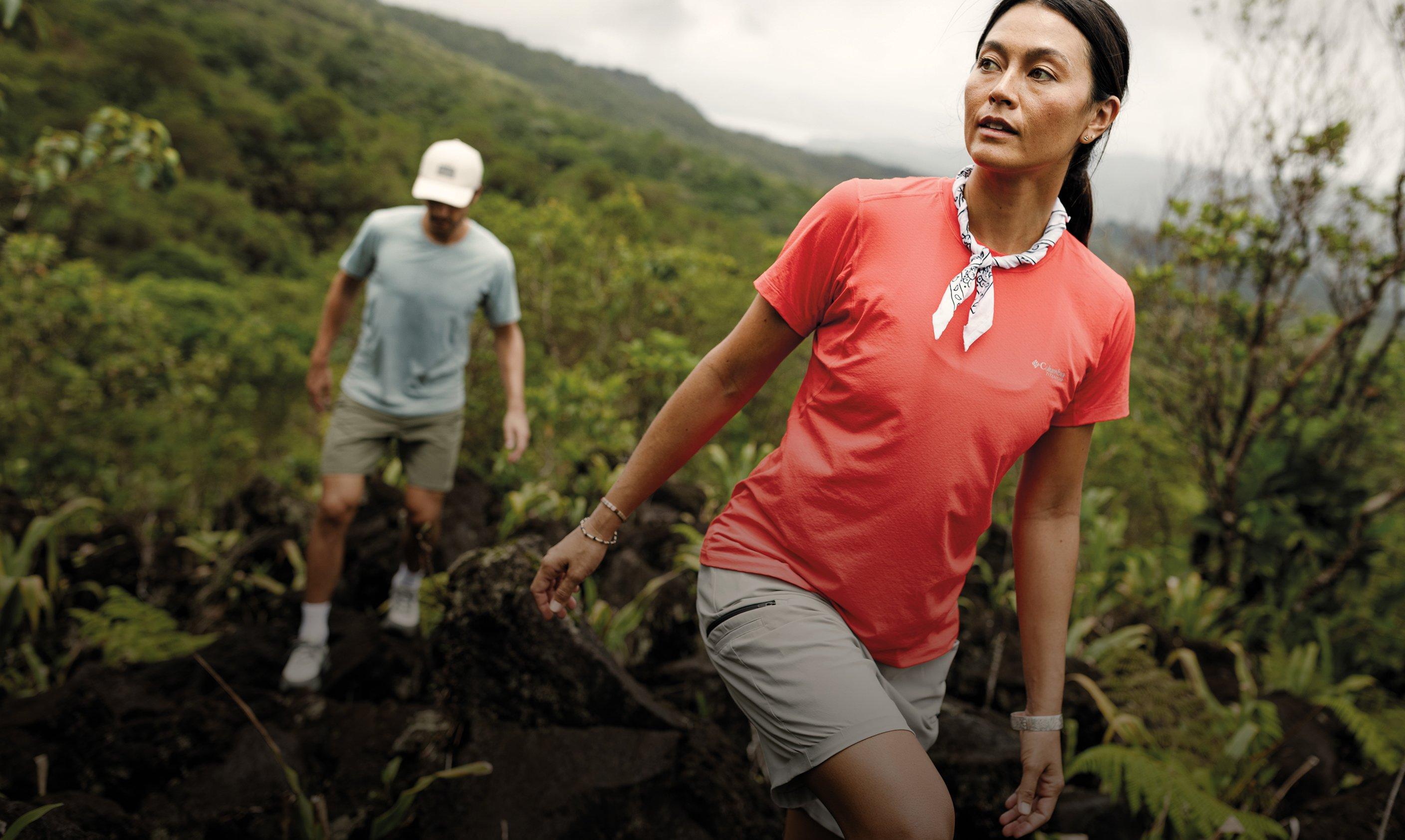 Woman hiking in coral t-shirt and gray shorts with patterned bandana, man in background on mountain trail.