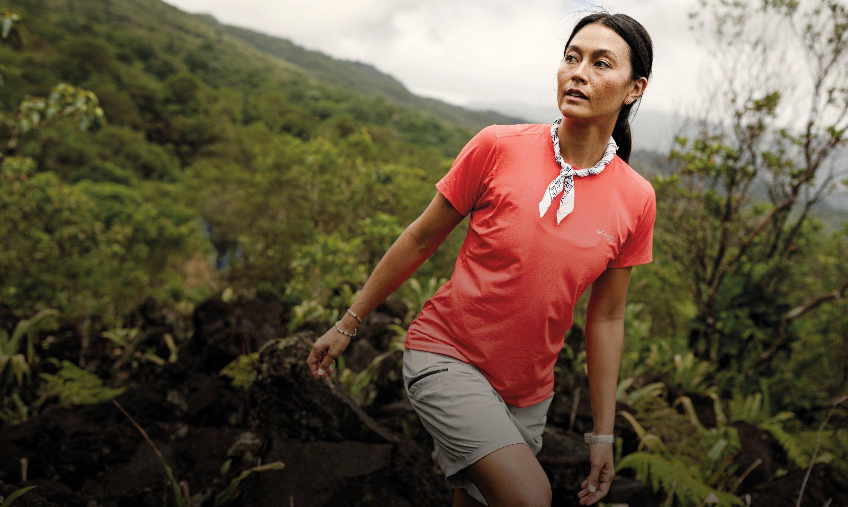 Woman hiking in coral t-shirt and gray shorts with patterned bandana on misty mountain trail.