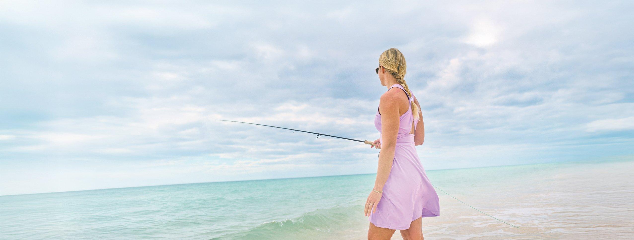 Woman in purple dress fishing from beach with rod, standing at water's edge under cloudy sky.
