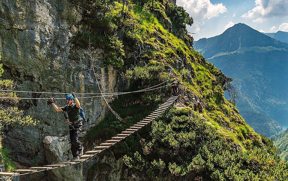 A person walking across a swinging wood and cable suspension bridge high above the ground in rocky, mountainous terrain. The bridge is a part of a via ferrata system and the person is safely attached to the cable so there’s no fear of falling.