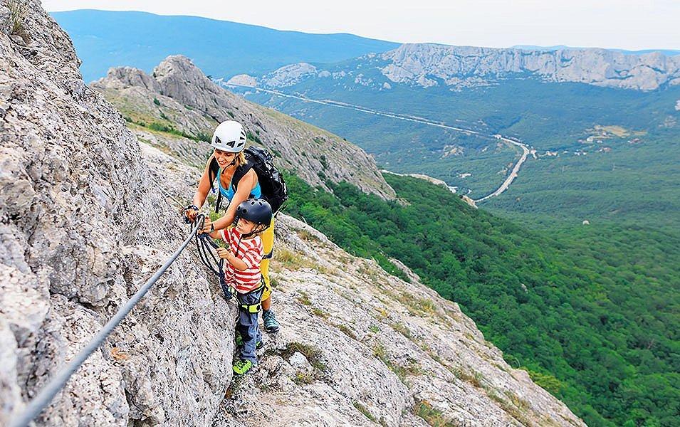 A woman and a child traversing a low-angled rock band on a rocky ridge high above the valley floor while attached to a safety cable that protects their progress.