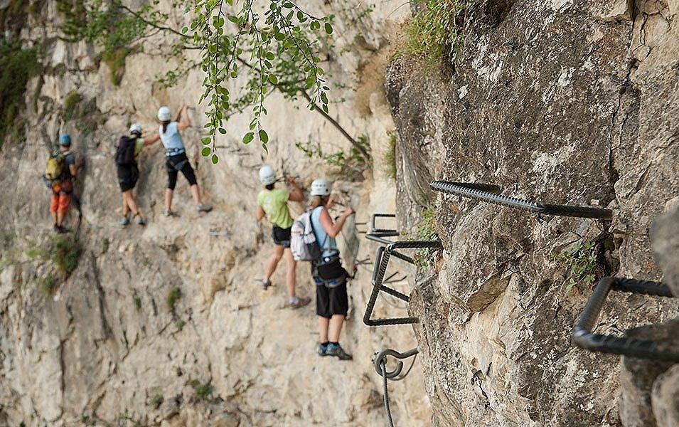 Five people appear to be climbing side by side on a steep rock face but closer inspection reveals that they are using metal hand and footholds and a safety cable that creates an easy path along the cliff.  

 