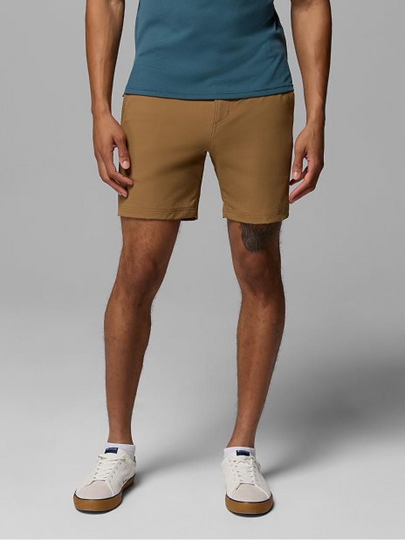Man wearing tan chino shorts with a teal shirt and white sneakers standing against a gray background.
