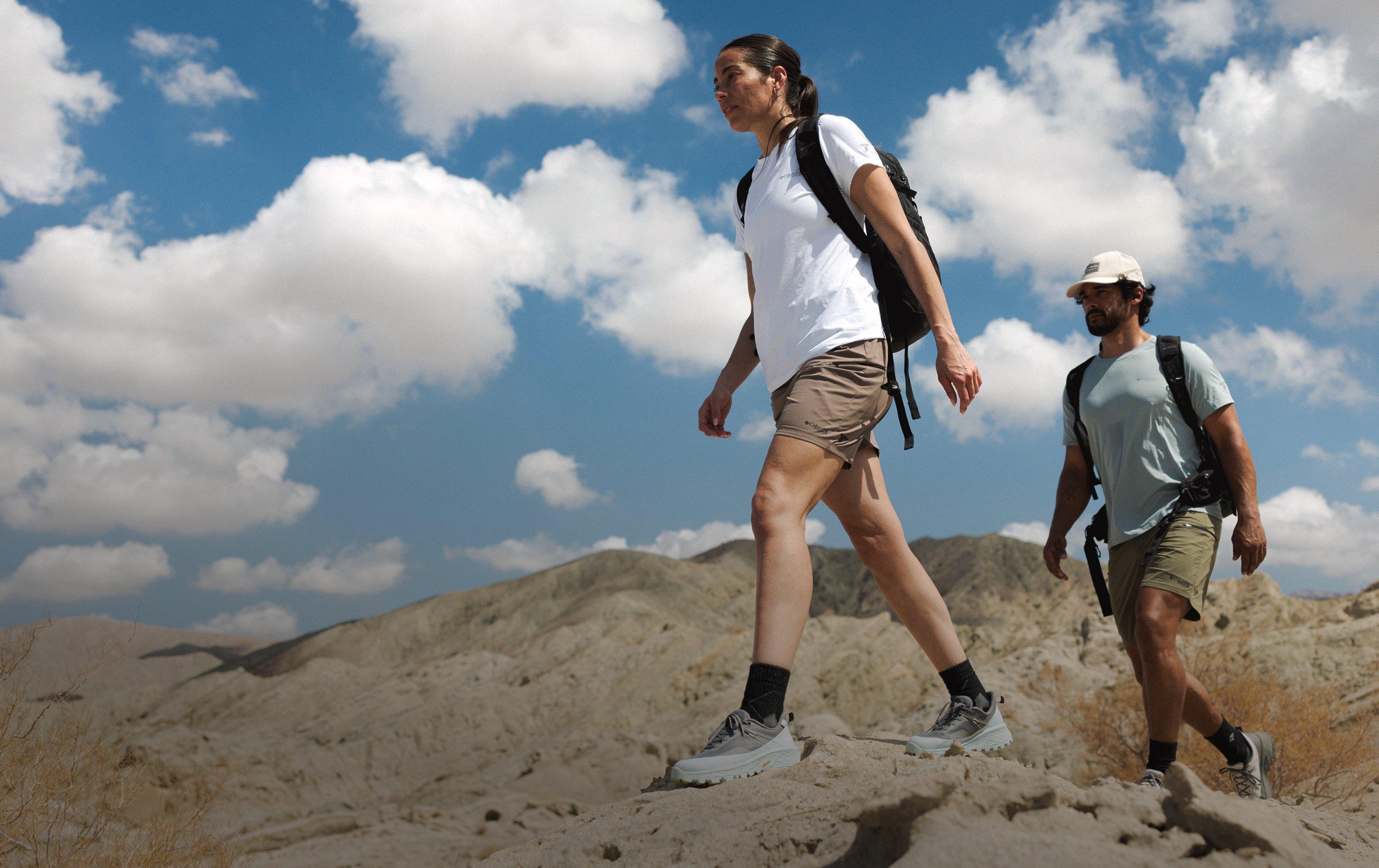 Two hikers with backpacks walking on desert terrain under a blue sky with white clouds.