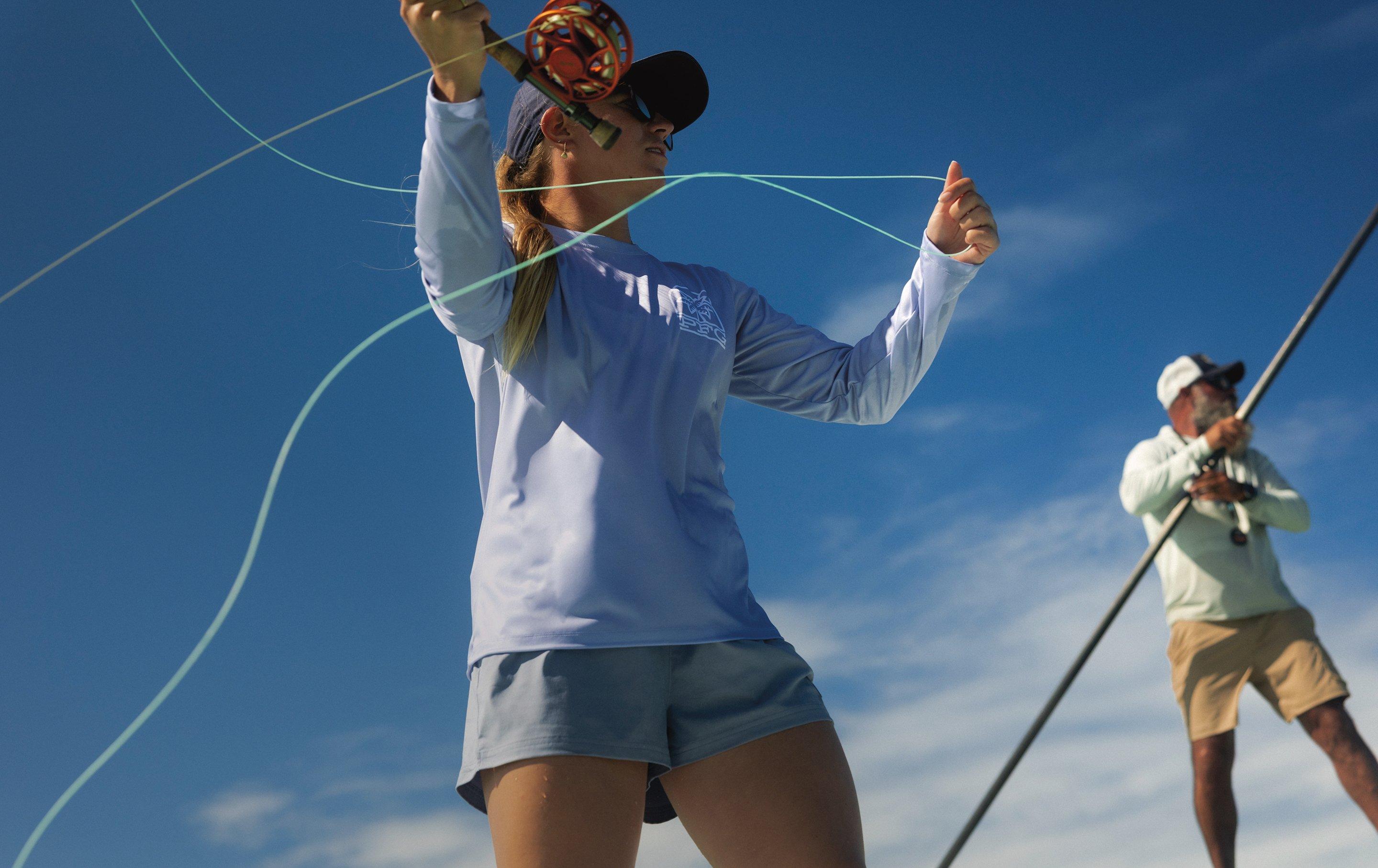 Woman in blue long-sleeve shirt and cap casting a fly fishing rod with another angler in the background.