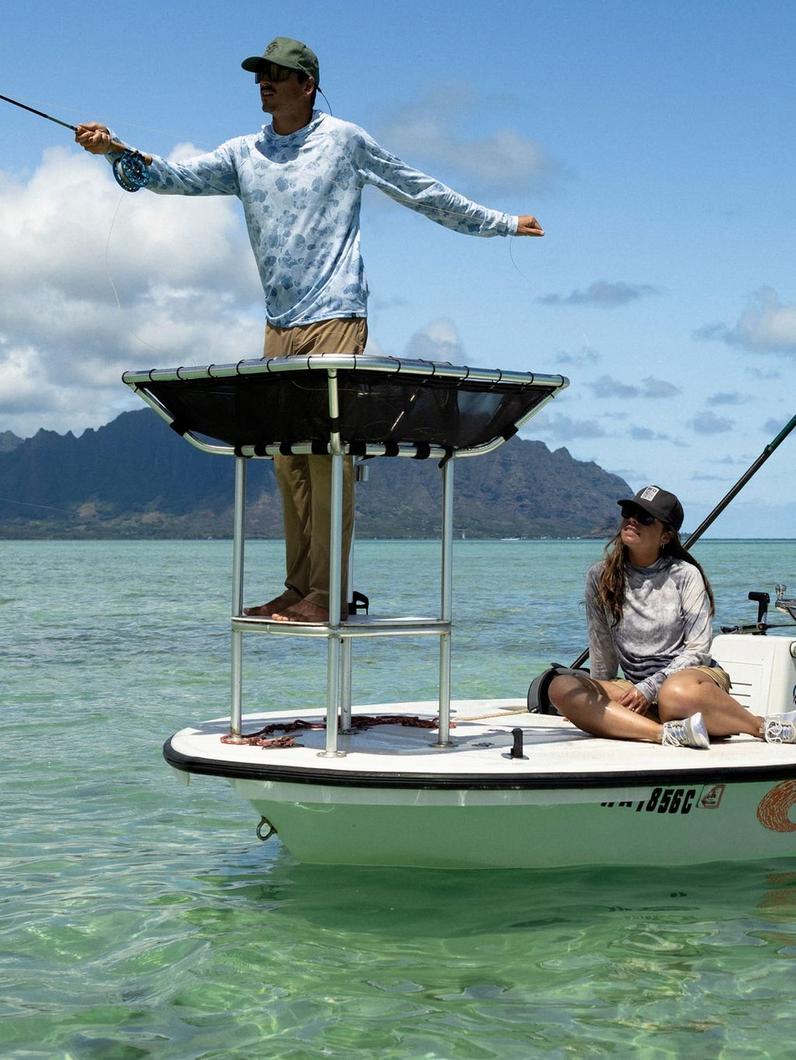 Man in blue fish-print long sleeve shirt and cap casting fishing rod from elevated platform on boat.