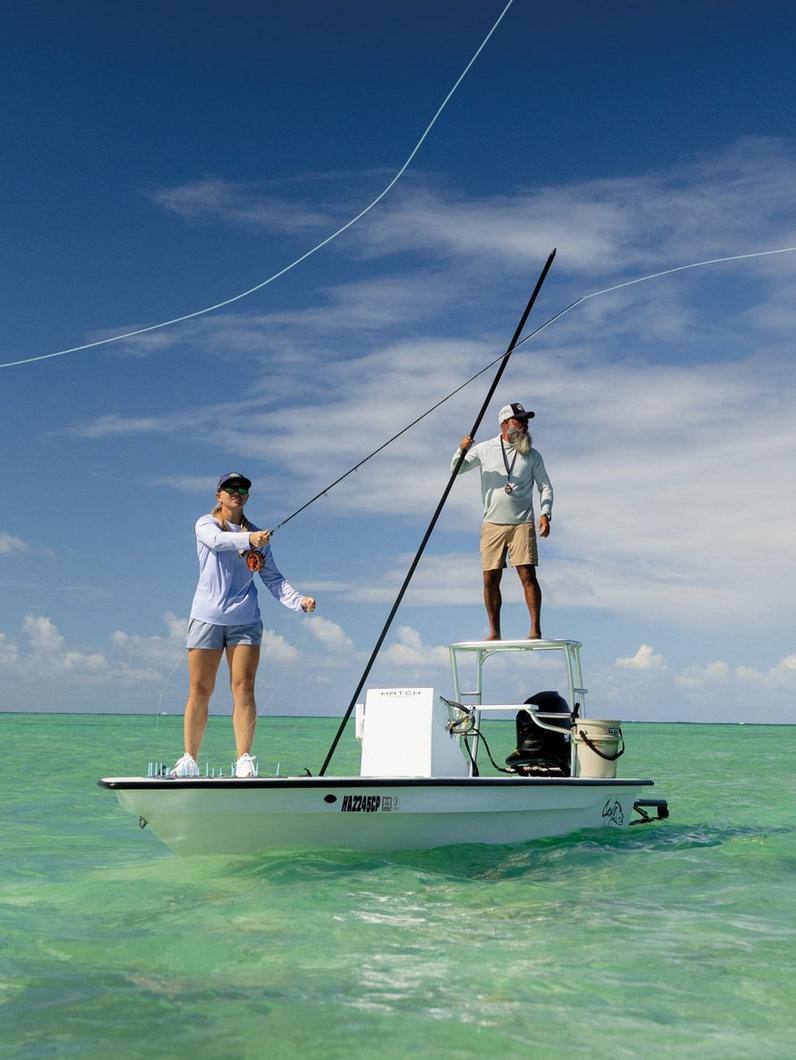 Two people fishing from a flats boat in shallow turquoise water under a blue sky.