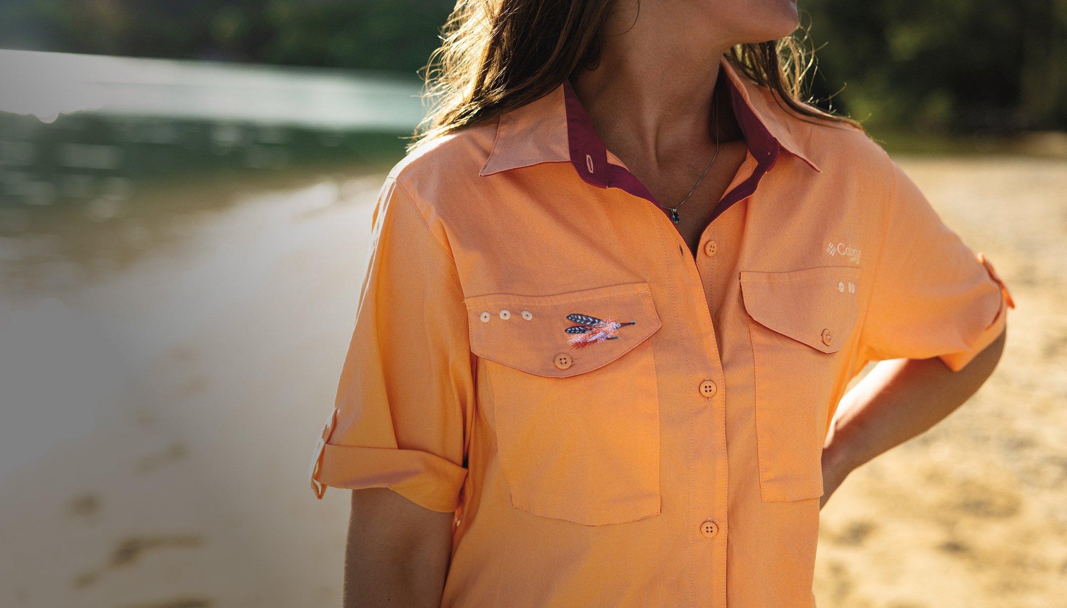 Woman wearing orange button-up fishing shirt with roll-tab sleeves and embroidered flag fish detail on pocket.