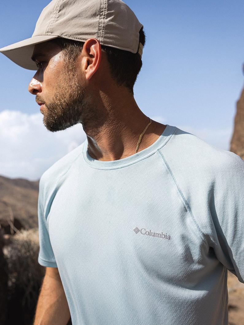 Man wearing light blue performance t-shirt and tan cap outdoors in natural setting.