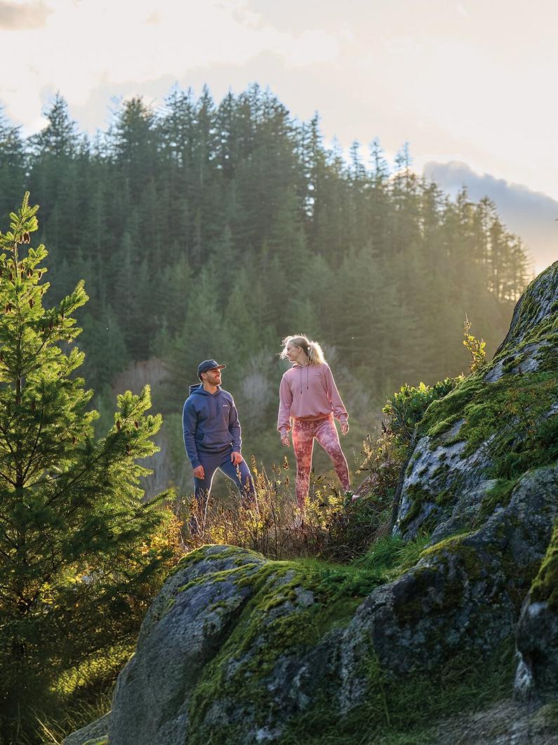 Two people hiking on a rocky, moss-covered trail surrounded by evergreen trees in natural sunlight.