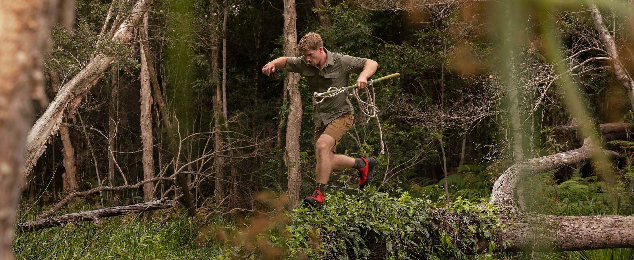 Man jumping over fallen log while trail running through forest wearing olive shirt and red athletic shoes.