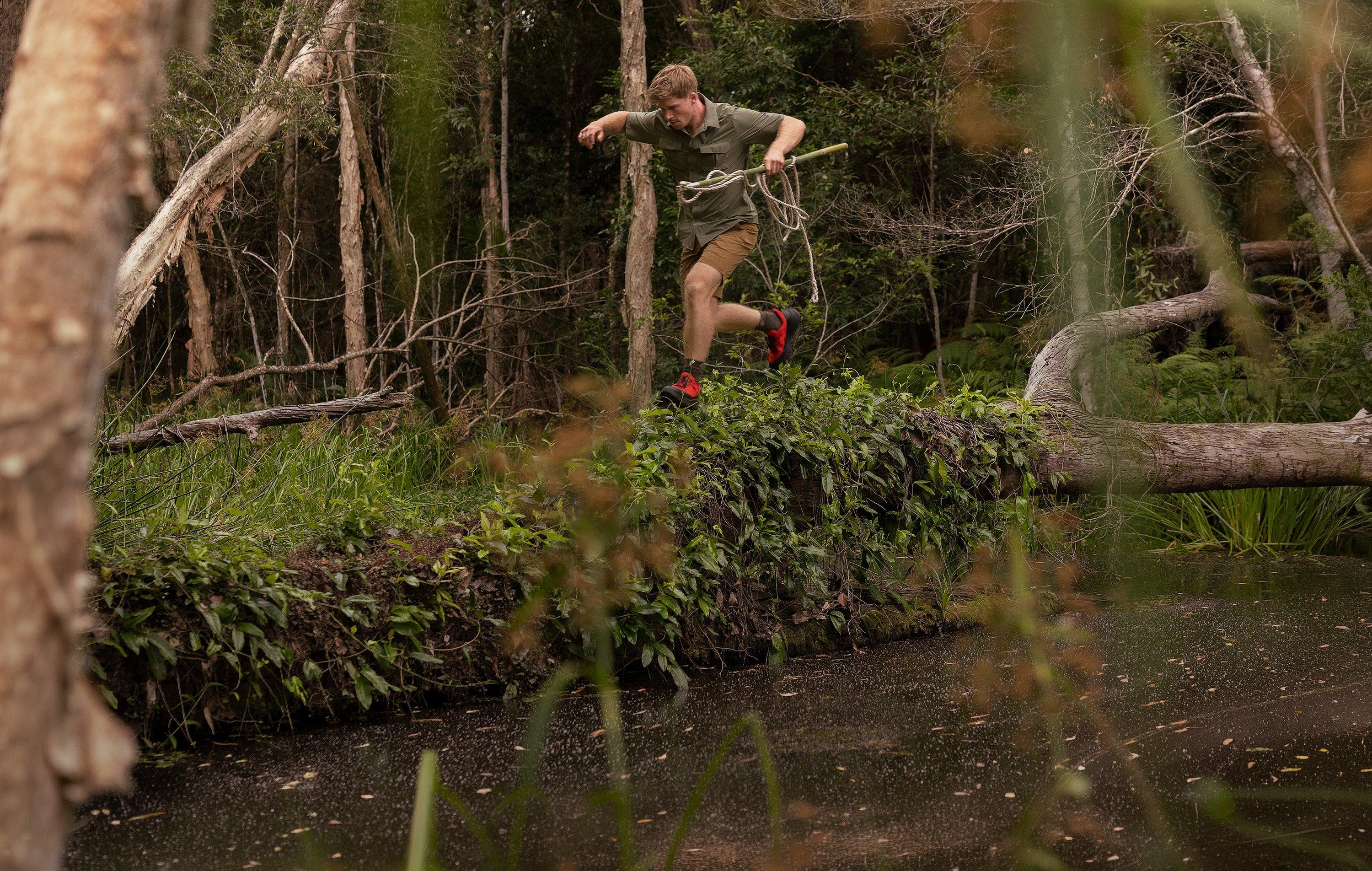 Man trail running through forest, jumping over fallen log wearing olive shirt, tan shorts, and red shoes.