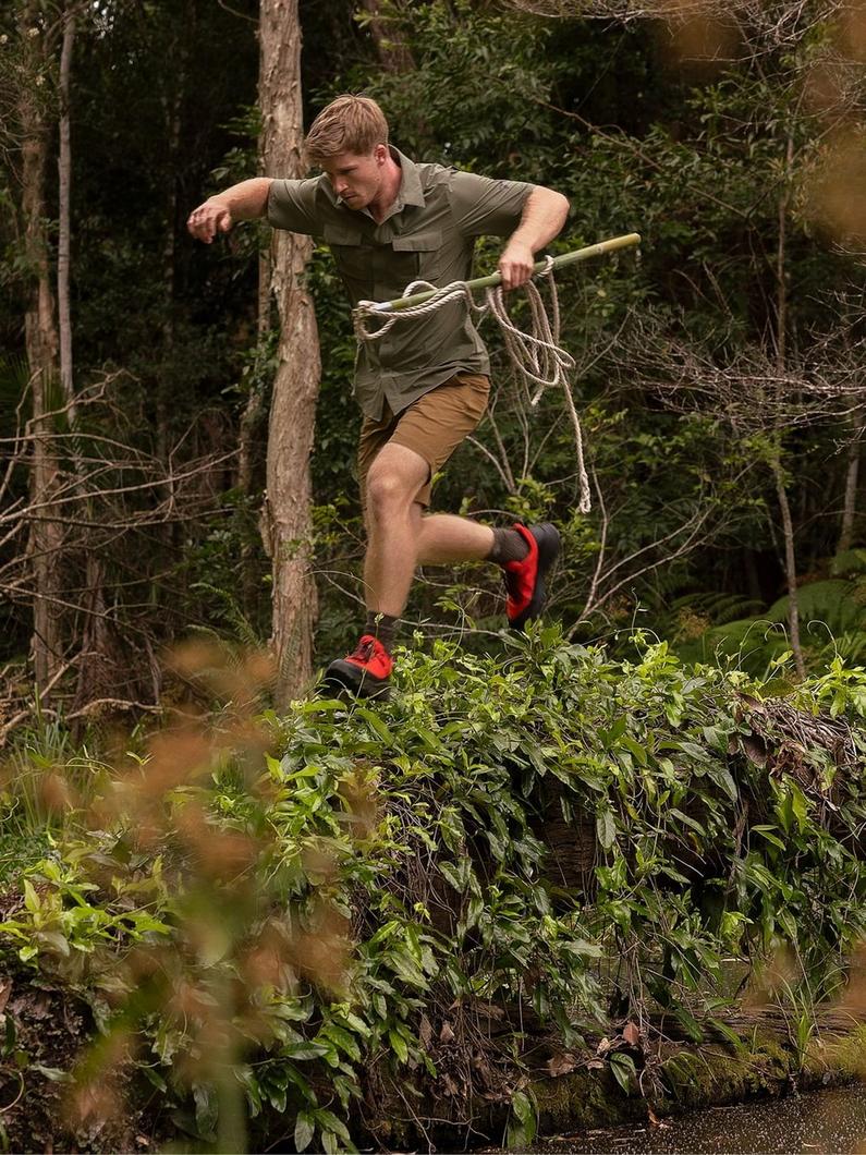 Man in olive shirt and tan shorts jumping over obstacle while holding rope in outdoor forest setting.