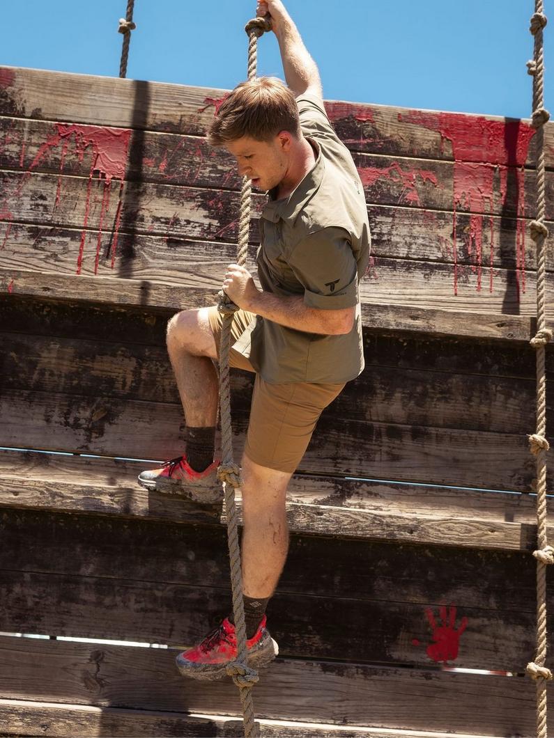 Man climbing rope obstacle on weathered wooden wall during outdoor fitness course.
