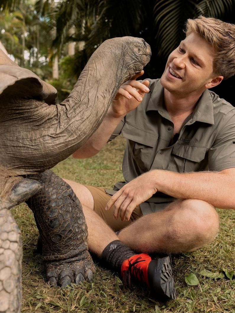 Man in khaki shirt smiling while gently touching a giant tortoise's neck in a tropical setting.