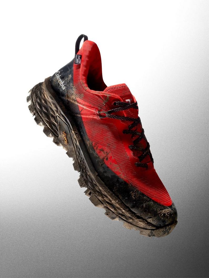 Red trail running shoe with black laces and aggressive tread, shown covered in mud.