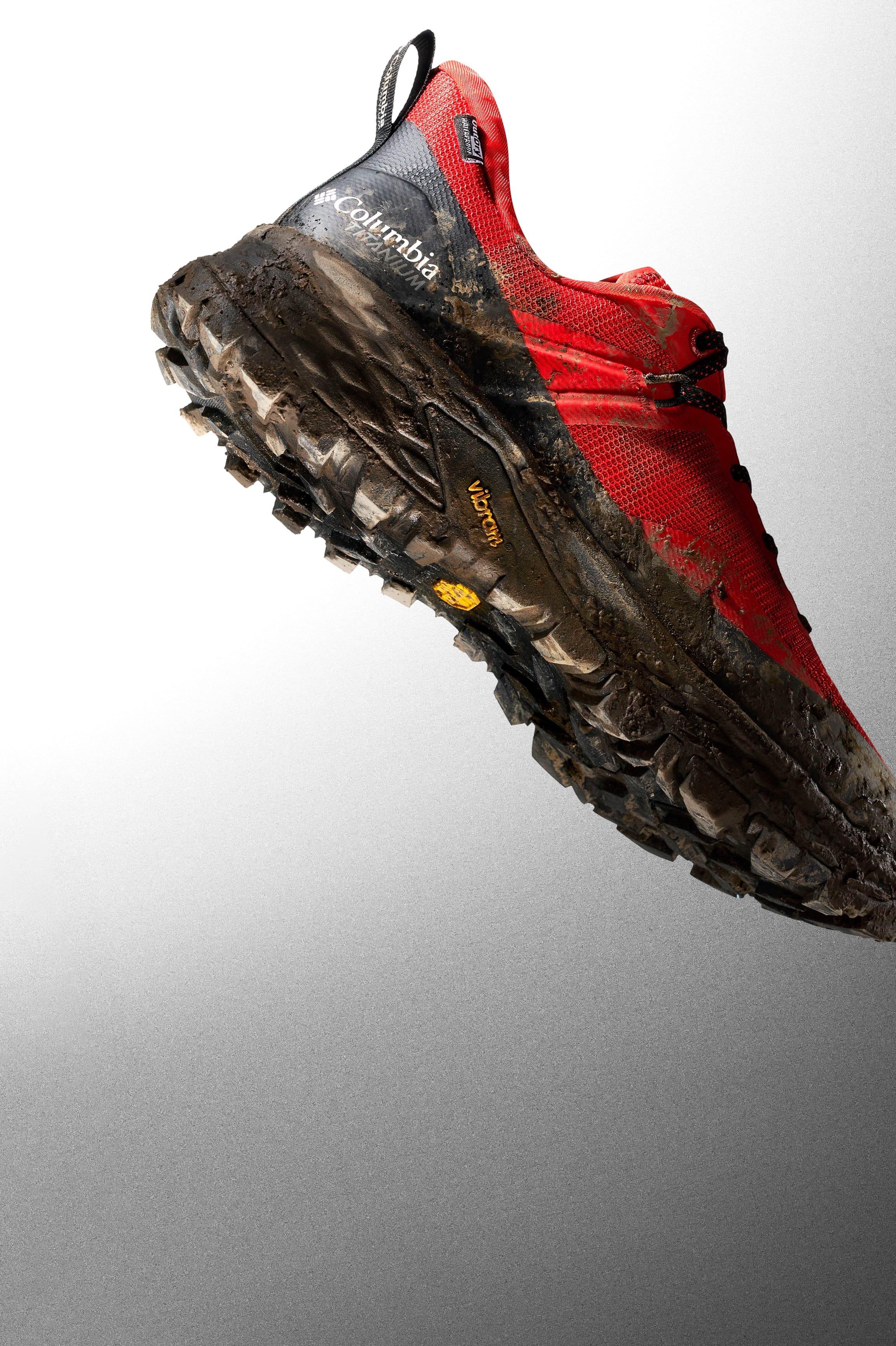 Red and gray trail hiking shoe with muddy, aggressive tread sole shown at an angle.