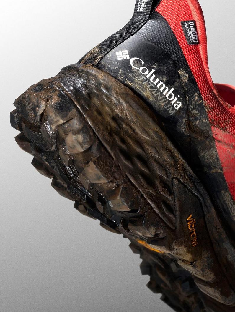 Close-up of muddy hiking boot sole showing deep tread pattern and red mesh upper panel.